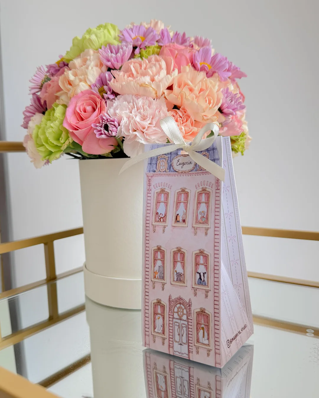 Cajita de papel en forma de edificio rosa, junto a un arreglo de flores