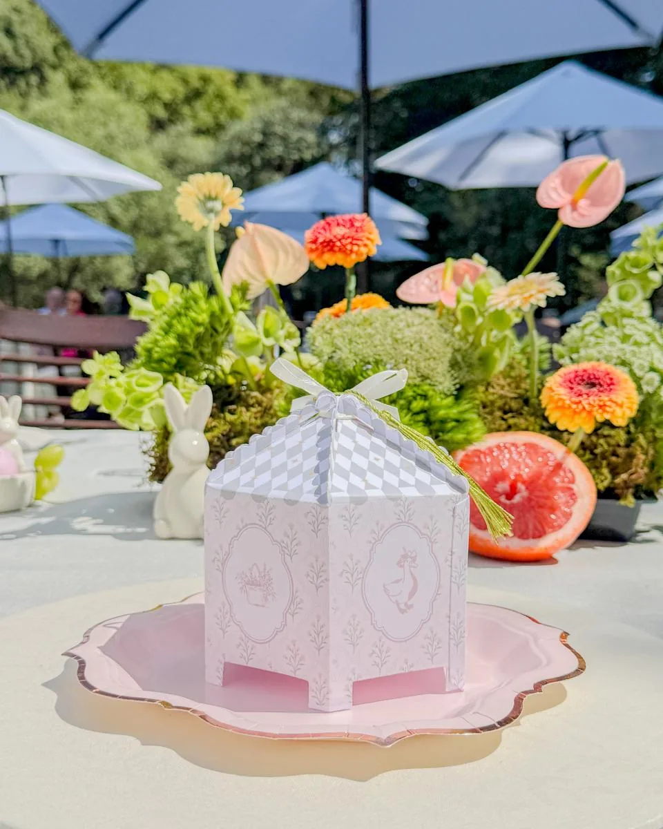 Cajita de papel en forma de carrusel sobre un plato rosa, en una mesa de jardín con flores