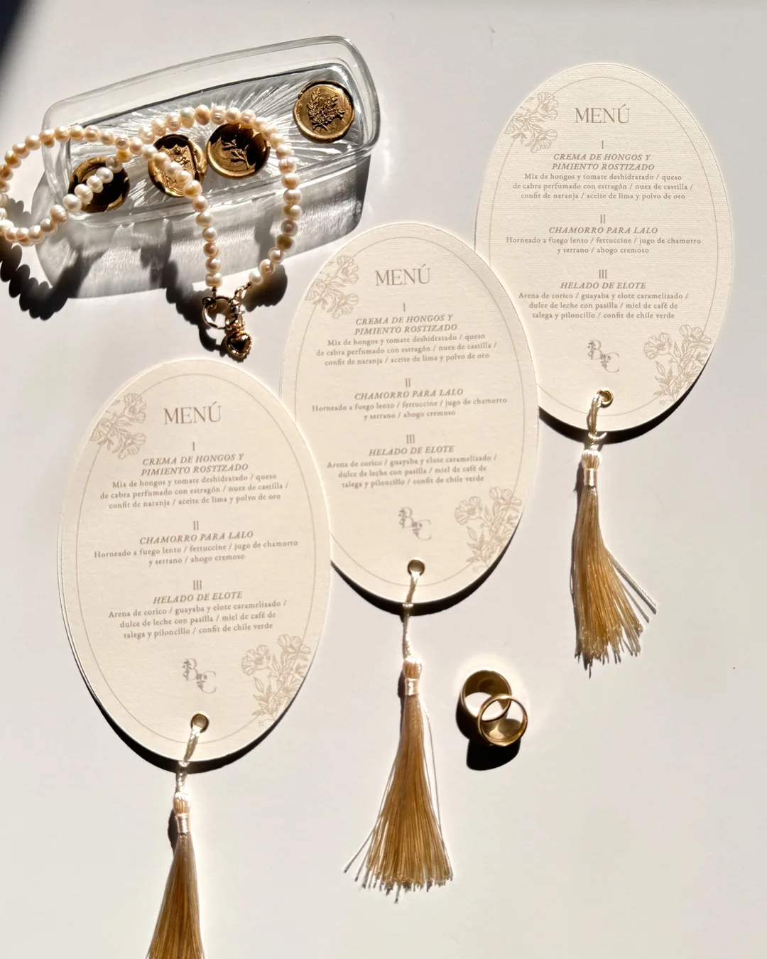 Tres menús ovalados color crema con borla dorada, junto a perlas y anillos